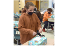A Tioga Hills Elementary fifth-grade girl wearing a brown hoodie and black mask applies one last piece of tape to the package she is wrapping in holiday paper.