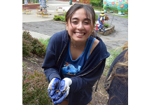 A Vestal High School senior girl in blue hoodie and wearing gardening gloves carefully cradles a tiny baby toad in her hands.