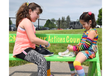 A Tioga Hills Elementary fifth-grade girl sits on a green bench facing a dark-haired Kindergarten girl, each with their sneakers in front of them, as the older girl teaches the younger one how to tie her shoes.