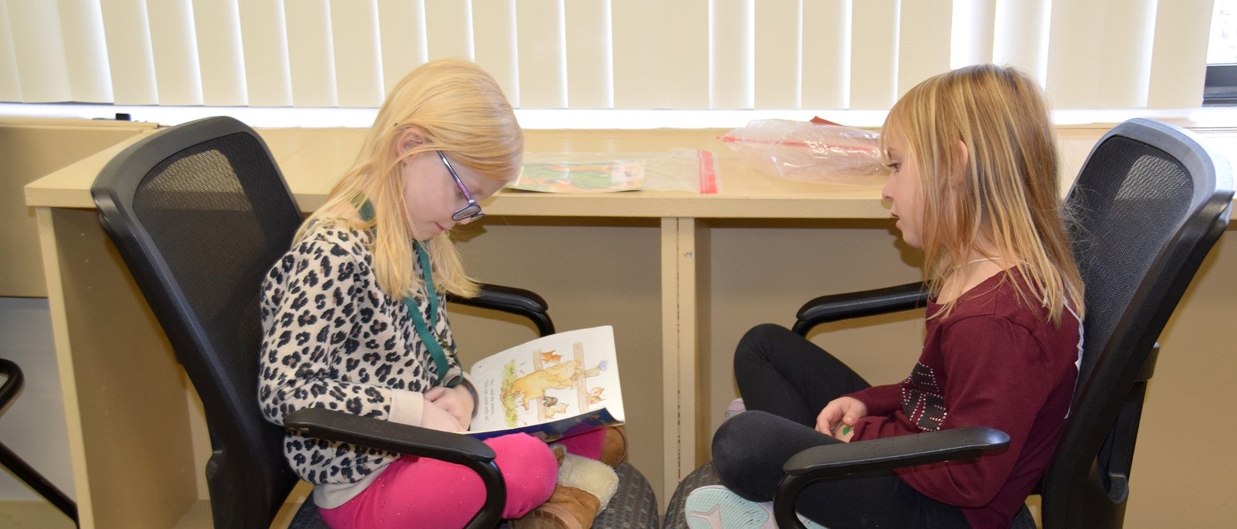 Two Glenwood first-grade girls sit in chairs facing one another as one reads to the other.