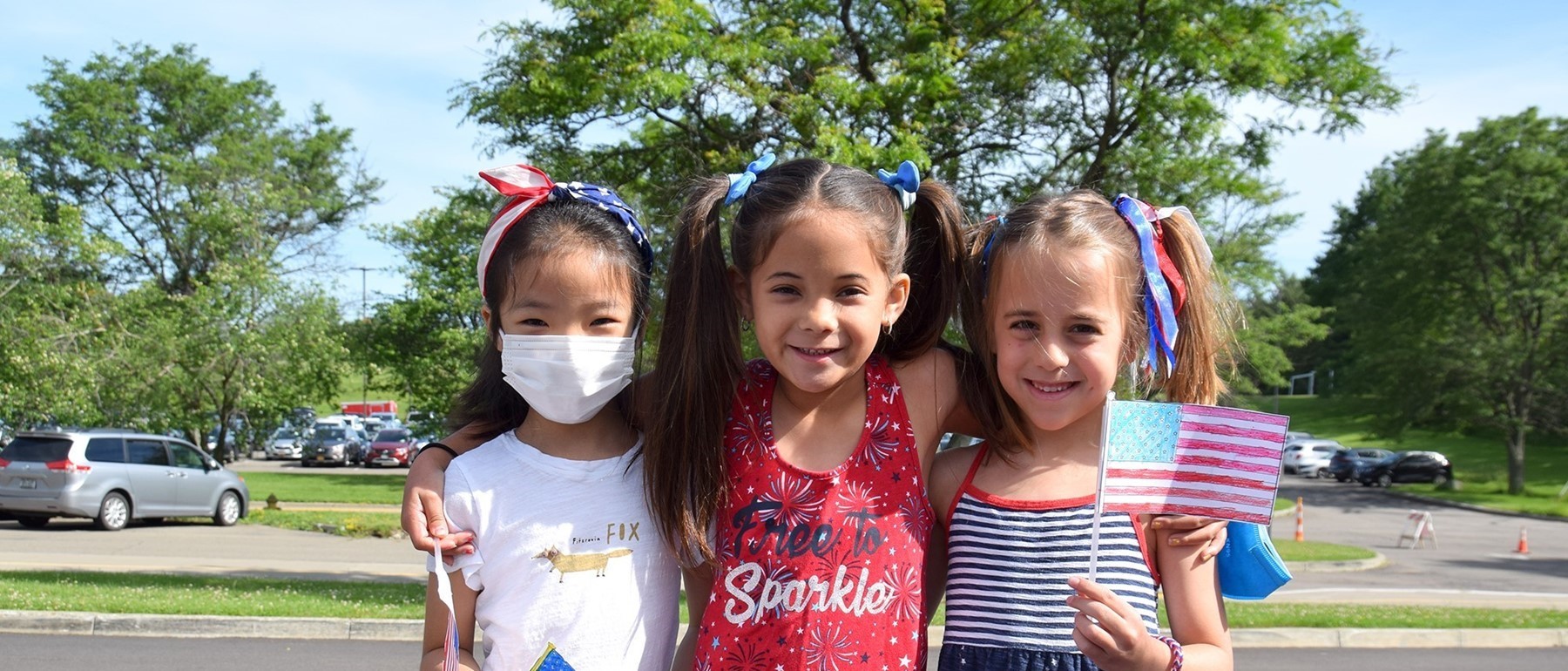 Three little girls from African Road Elementary are dressed in red, white and blue attire and accessories and two hold paper flags on popsicle sticks.
