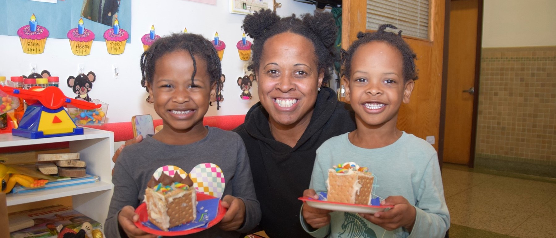 Twin Universal Pre-Kindergarten students at the Cub Care campus proudly hold their decorated gingerbread houses as they stand on each side of their mother.